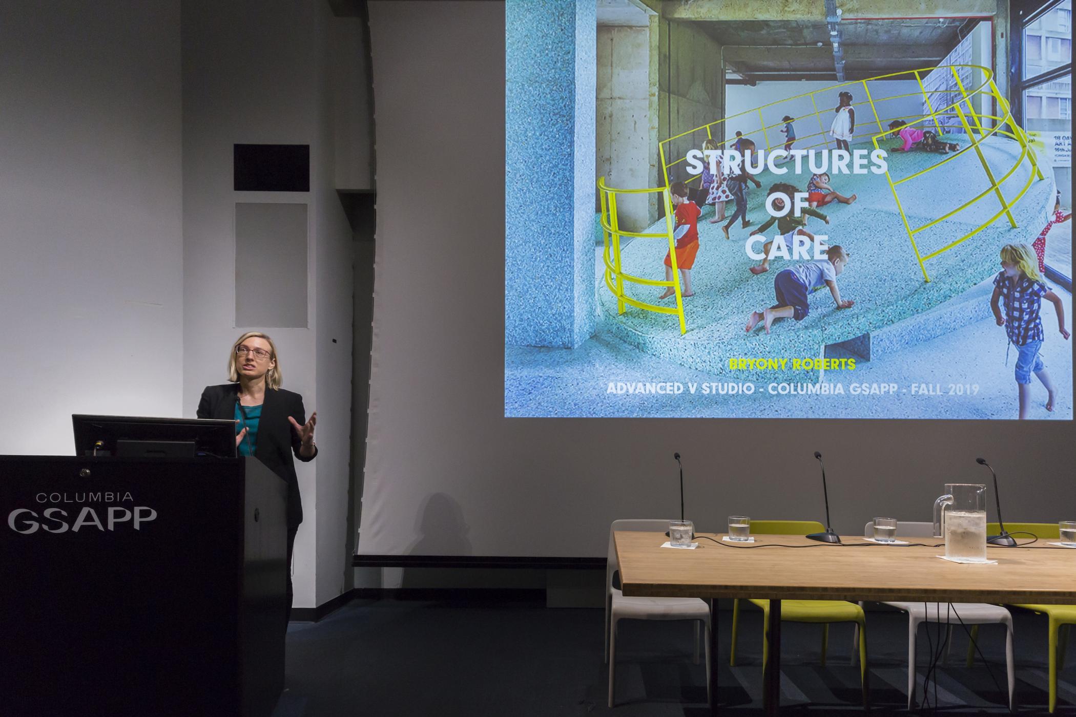 Bryony Roberts stands at a podium gesticulating beside a slide showing children playing on a tilted platform with text saying "structures of care"