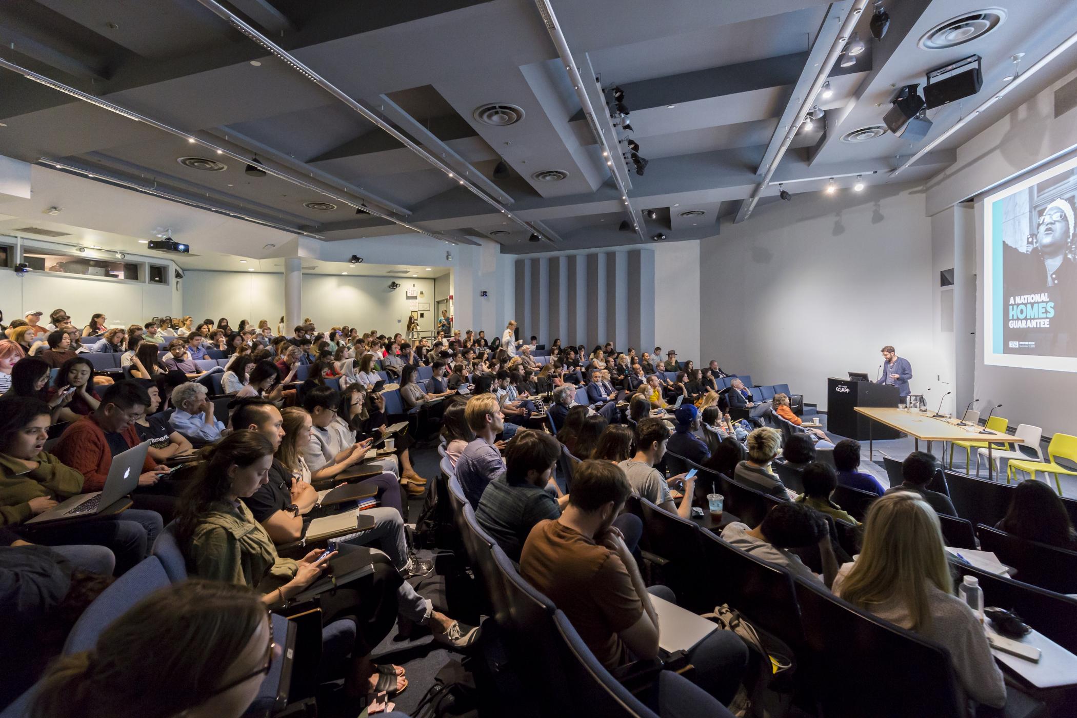 Wide angle view of a full lecture hall with audience watching Daniel Aldana Cohen present in front of a slideshow