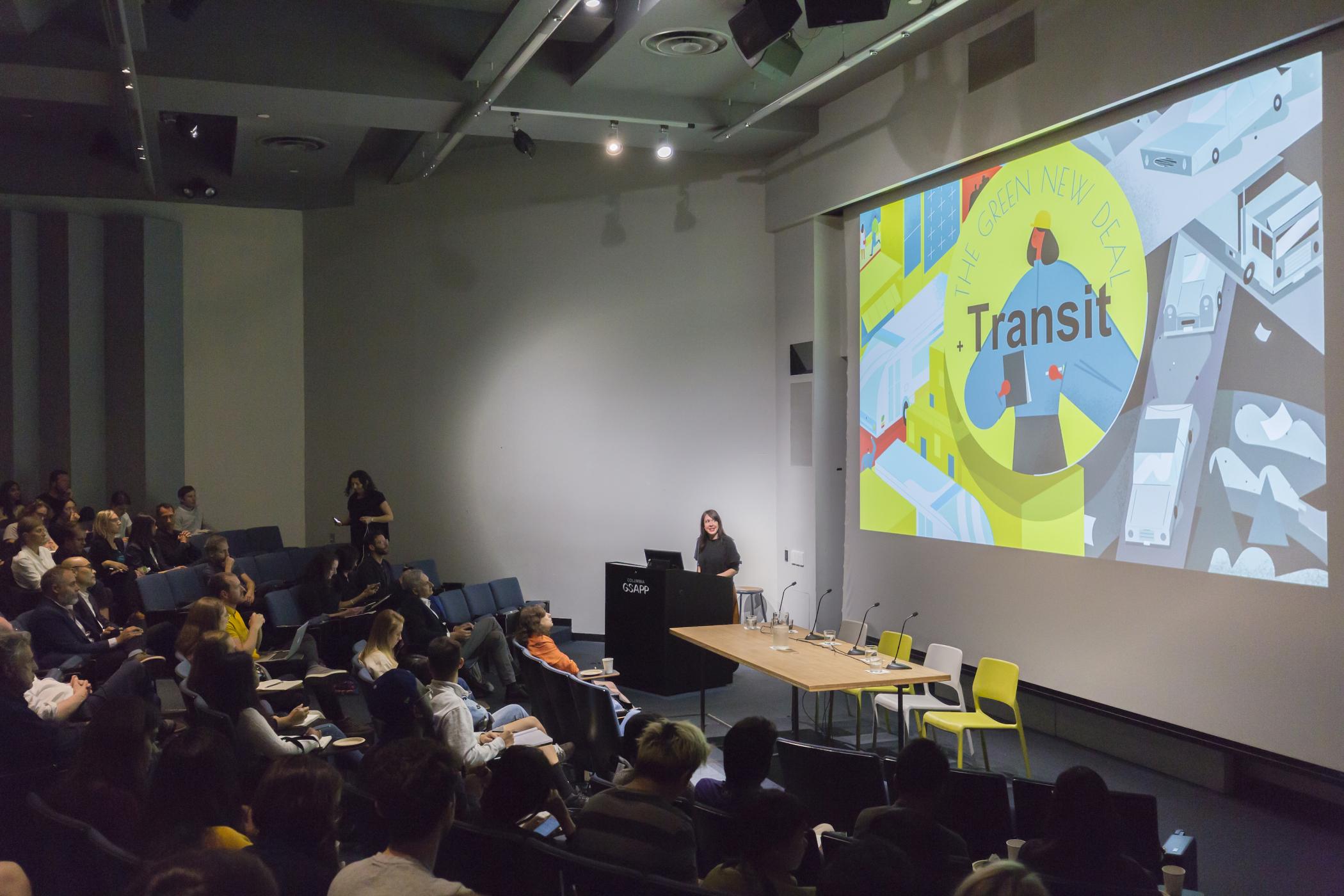 A wide-angle shot of Hayley Richardson behind the podium next to a slide with text saying "transit" in front of graphic design