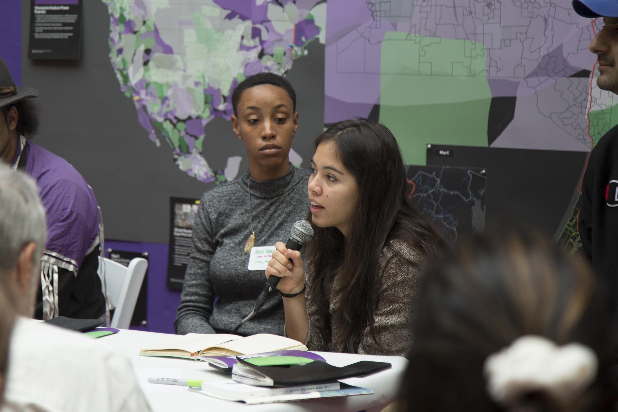 Xiye Bastida holds a microphone to speak as part of the "Democracy and Power" workshop while another participant watches her in front of a purple and green map of North America