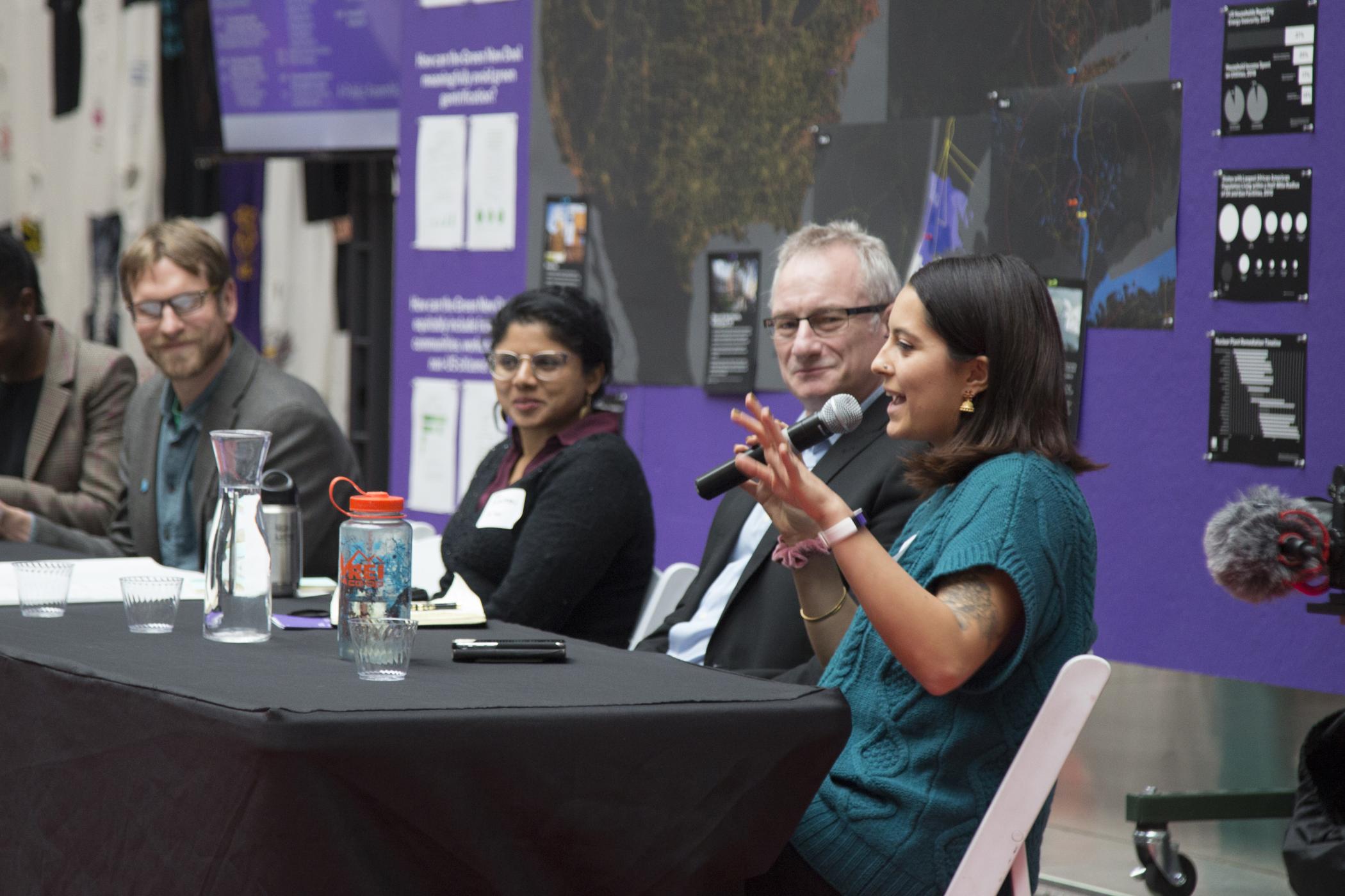 Priya Mulgaonkar gesticulates while holding a microphone sitting at a black table next to four other panelists during the "Energy and Power" discussion